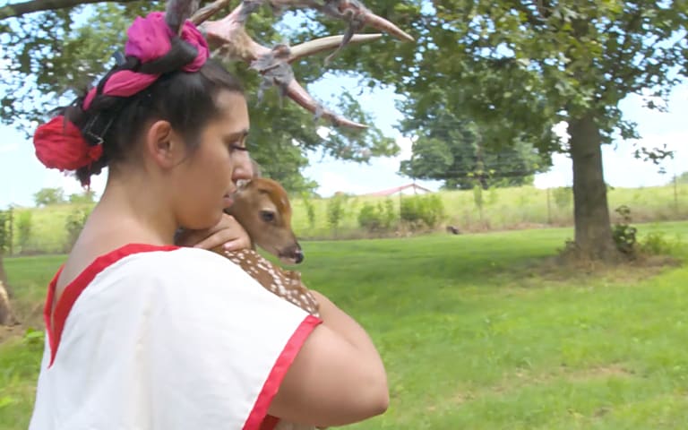 Video still of a woman holding a deer as she walks through a farm-like scene.