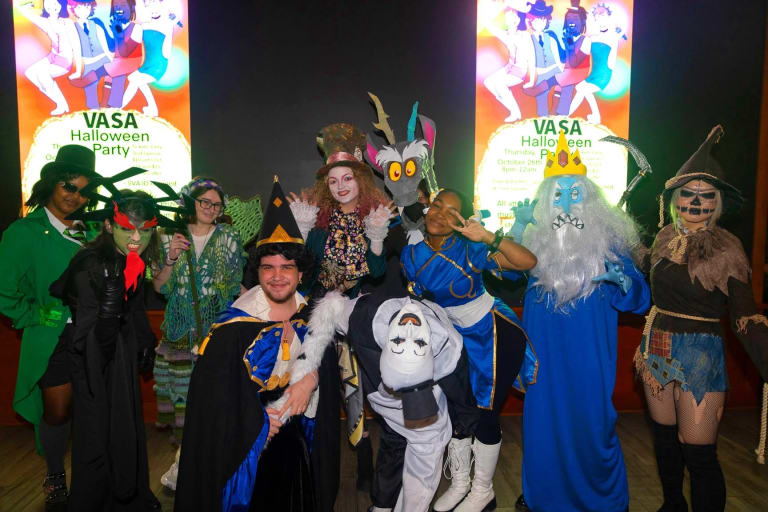 Students in colorful Halloween costumes standing in front of a sign that reads 'VASA Halloween Party."