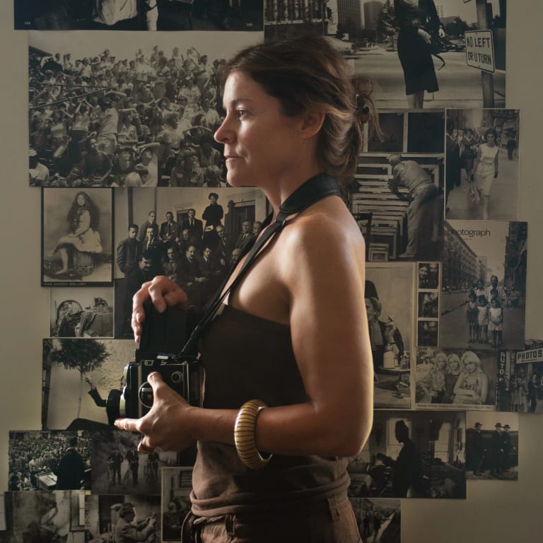 A photograph of a woman in profile facing left as she holds a Rolleiflex camera in front of a background of many black-and-white-images taped to a wall