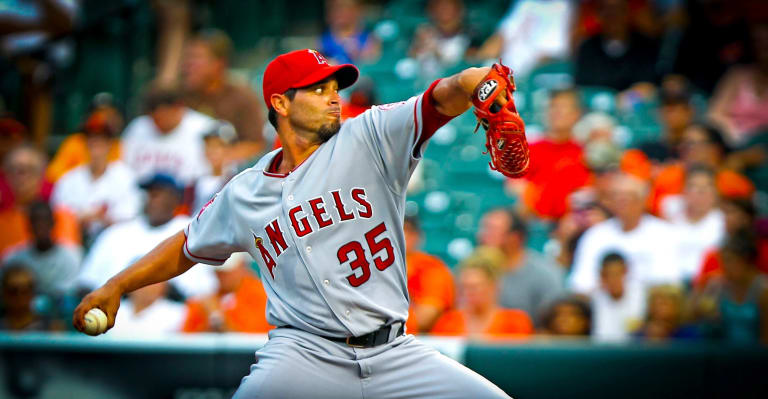 A man in a Los Angeles Angels jersey with he arm wound up to pitch a baseball. the crowd behind him is out of focus.