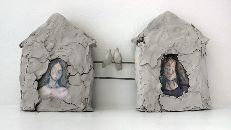 Clay sculpture of two house, each containing an abstract painting of a woman, with the houses linked by two metal lines. On the top metal line are two songbirds.