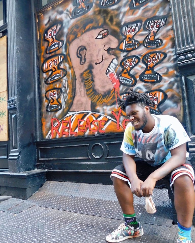A photograph of a man sitting in front of a building exterior decorated with a mural.