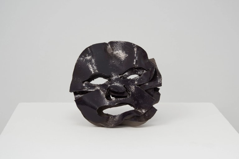 A very dark gray ceramic sculpture of a round mask made of simple shapes, sitting in a white pedestal