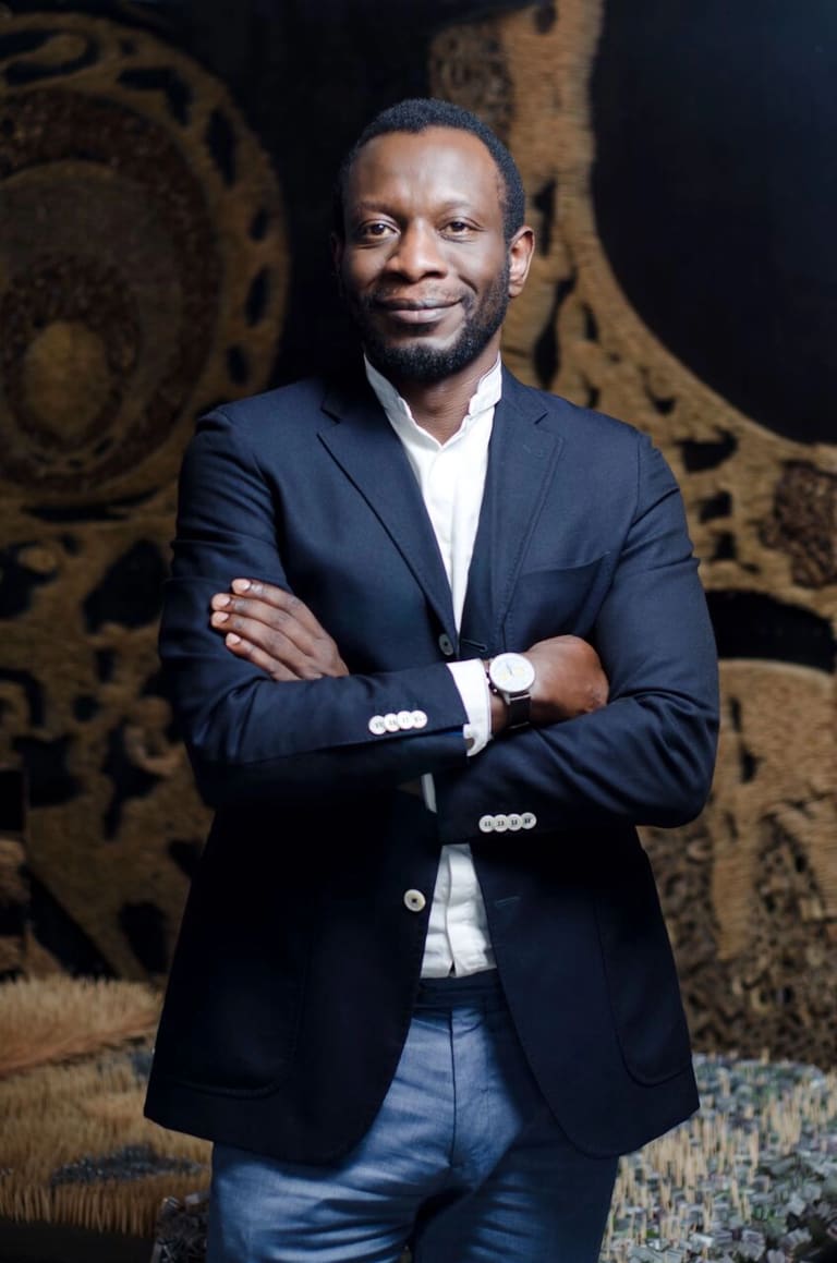 Color portrait of Azu Nwagbogu, wearing a white shirt, navy jacket and jeans. He's smiling and his arms are crossed. 