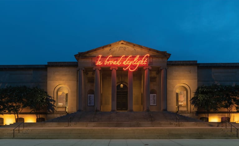 Installation view of Tavares Strachan’s work In Broad Daylight, installed at the Baltimore Museum of Art in 2018. A neoclassical style building at dusk features neon lights that read "In Broad Daylight" in reddish orange.