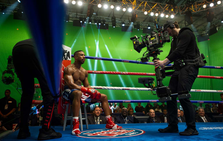 Color photograph of a cameraman using a Steadicam to record actor Michael B. Jordan on a boxing ring set built in a green screen studio.