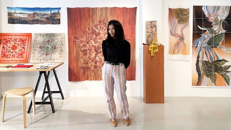 Woman in black turtleneck stands in front of wall with various tapestries