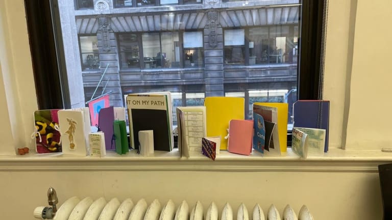 Picture of multiple handmade books in different colors, shapes, and sizes. They are standing on a white window sill, with a radiator underneath and a building in the background.