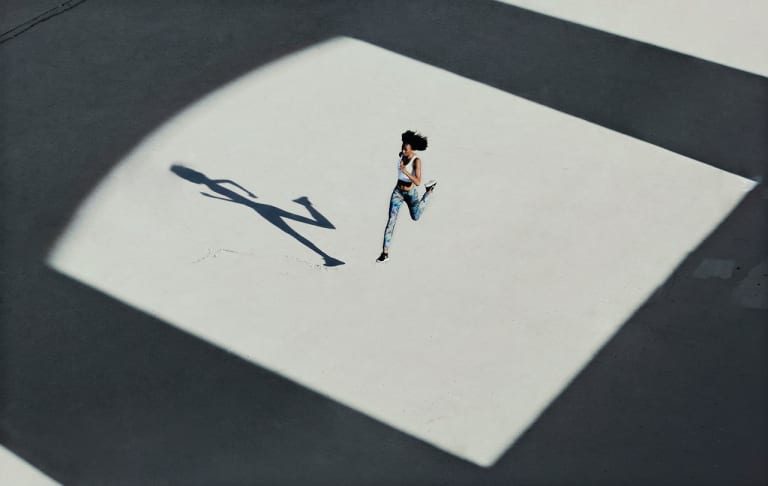Horizontal color photograph of a woman in athletic clothing running. She is photographed from above framed by a square of sunlight, surrounded by architectural shadows. She is mid-jump and we can see her shadow on the concrete floor