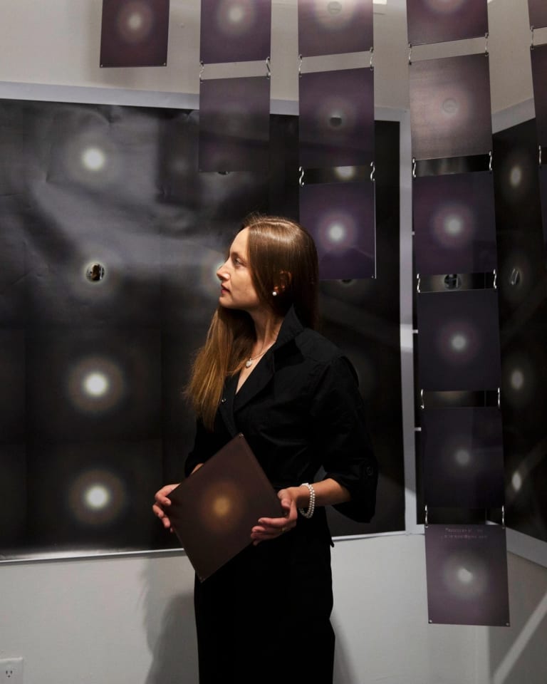 A woman stands in a dark artist studio surrounded by hanging black and white prints of the moon. She has long blondish brown hair and stairs off camera at her artwork.