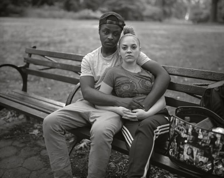 Black-and-white photograph of a man and a woman sitting on a park bench. She is leaning into him and he has his arms around her. He is looking away while she gazes directly into the camera. 