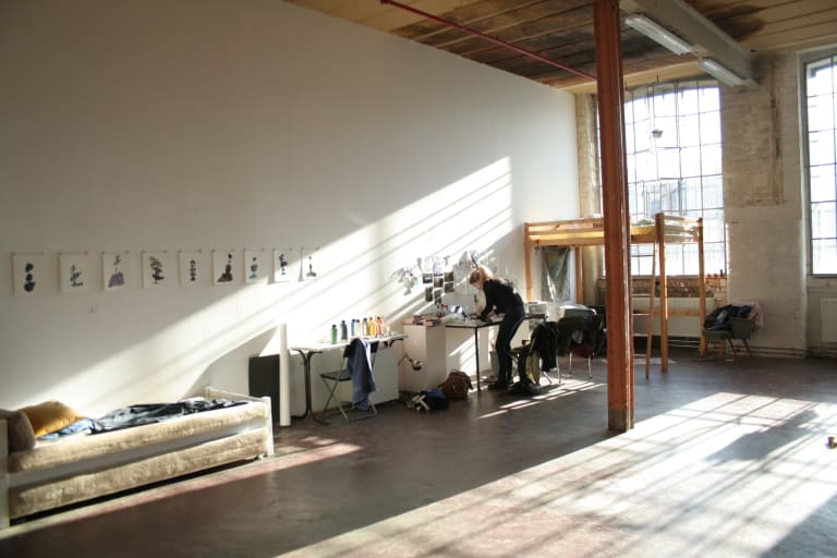 A photo of a large art studio with sunlight coming into the room from big windows in the furthest wall. To the left is another wall with a work table against it with a woman leaning over it. There is also a cough against the wall to the left, several black drawings on white paper taped to the wall, and a simple bunk bed in the furthest corner where the two walls meet.