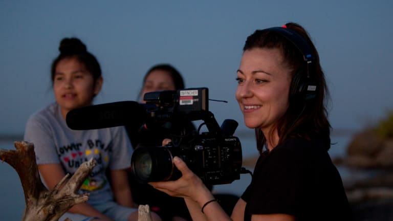 A smiling woman holds a videocamera, while standing outside. Behind her two young girls sit on a log.