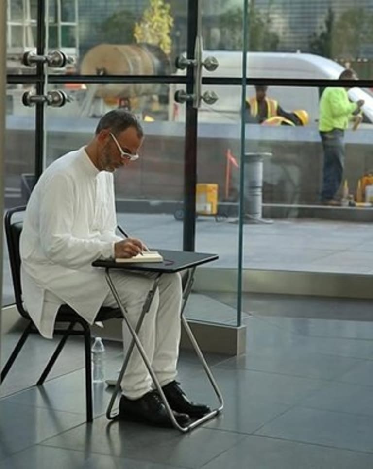 A picture of a seated man in middle eastern clothing writing in a book inside a glass building.