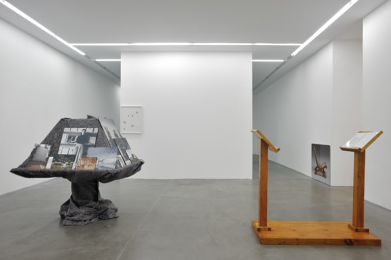 A mostly empty white room displaying four pieces of art. Two small wall displays and two standing objects.