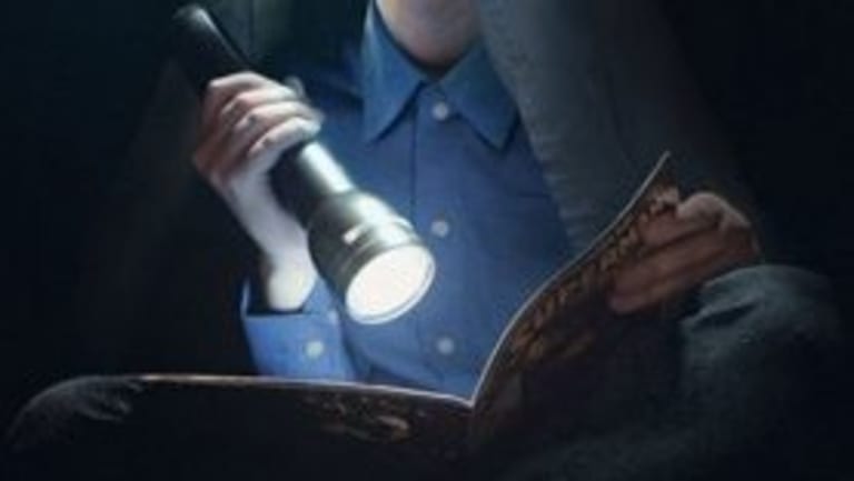 A person with a sheet over their head with a comic book with a flashlight. The person has bright glowing glasses staring in the distance. Below them is an object where you can only see the headlights.