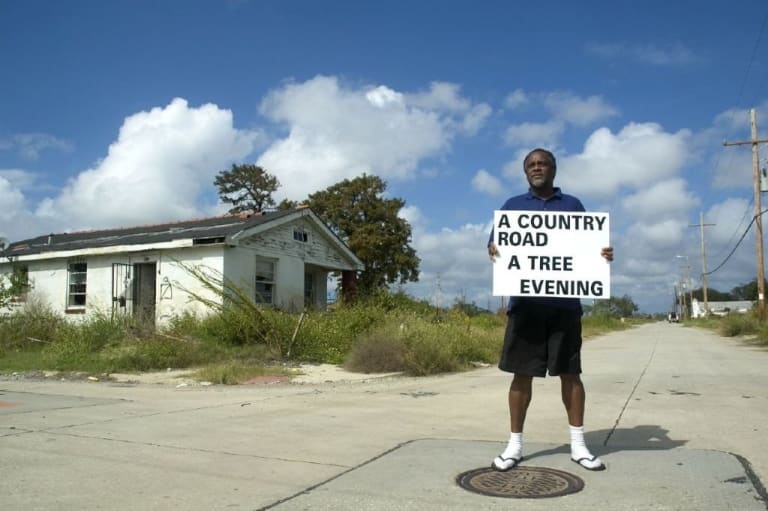 a man standing on the road holding a sign that reads a country road a tree evening