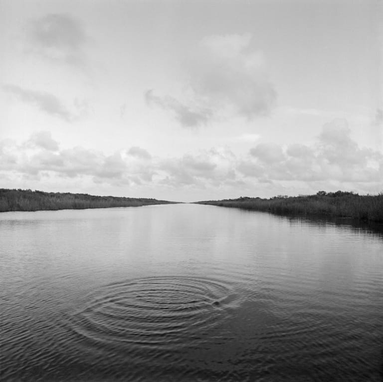 Black and white photo of a body of water expanding into the distance and meeting the cloudy sky at the horizon.
