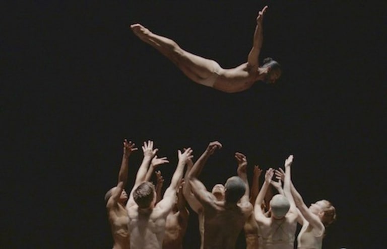 a group of men, dancers, stand shirtless in a circle, most with their backs to the camera. They toss another man, wearing only briefs, gracefully into the air