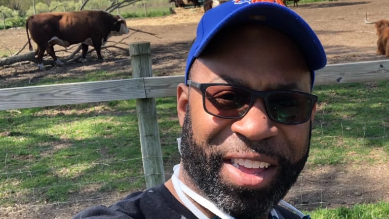 Here is a photo of Dr. Jarvis Watson standing outdoors at a farm, wearing a blue cap and dark sunglasses, a cow behind him. 