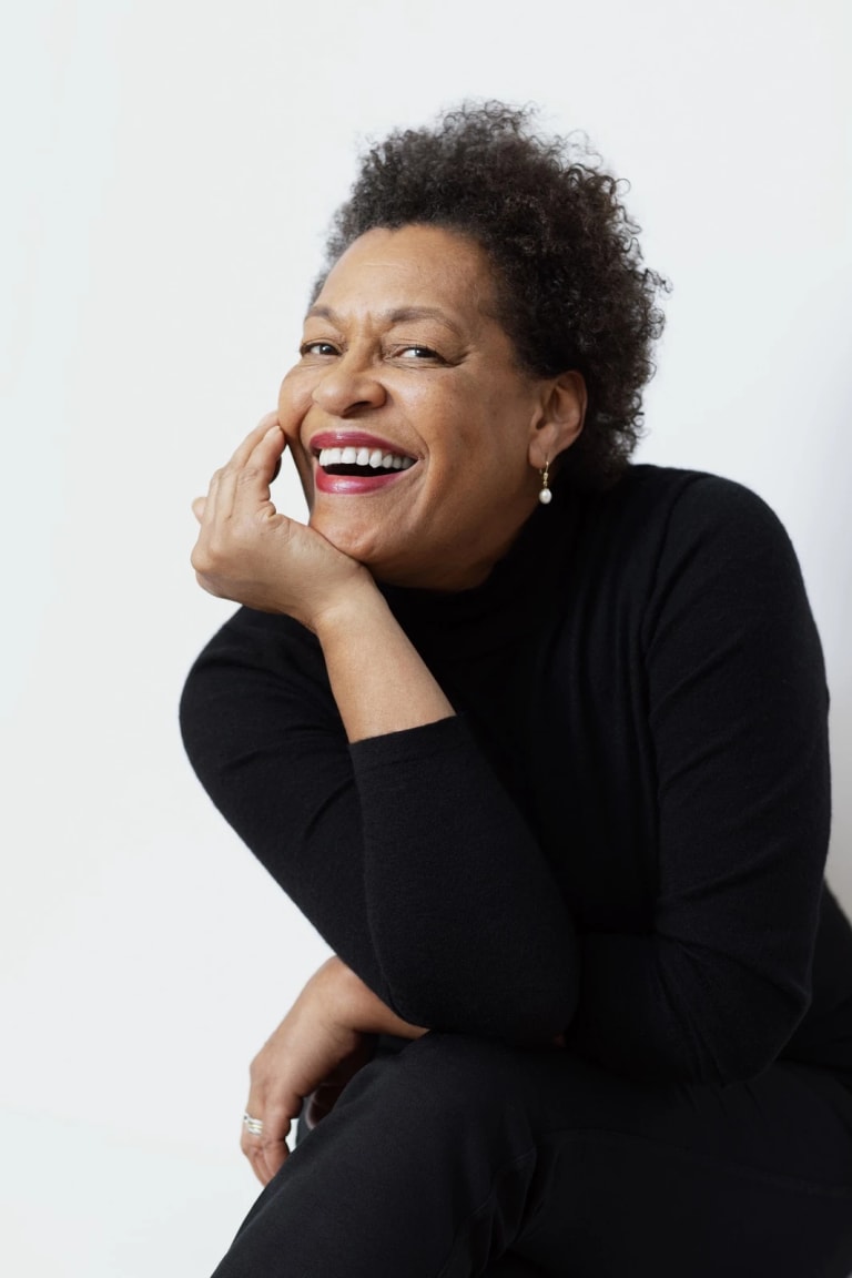 A black woman in a black turtleneck smiling in front of a white wall, resting her face in her hand