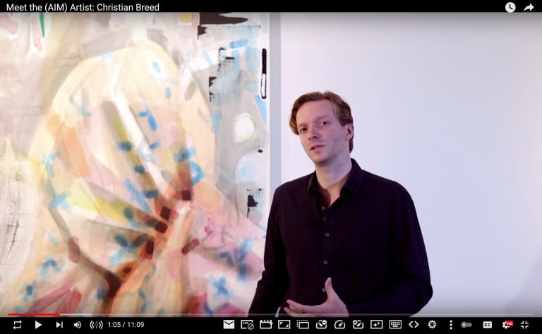 A video screenshot showing artist Christian Breed un a black long-sleeved shirt standing one front of one of his abstract paintings in a white museum space