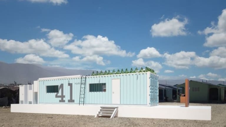 A pale blue shipping container that has been repurposed as a home sitting in the desert.