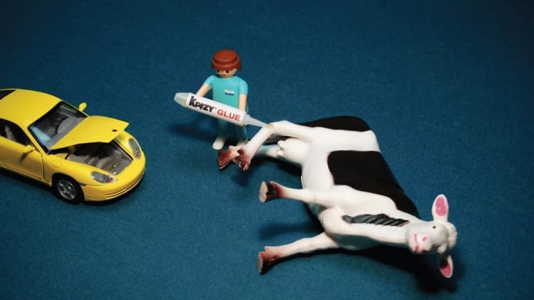 A yellow car with the hood popped open while a black and white cow lays on it's side getting it's leg glued by a man in a turquoise outfit.