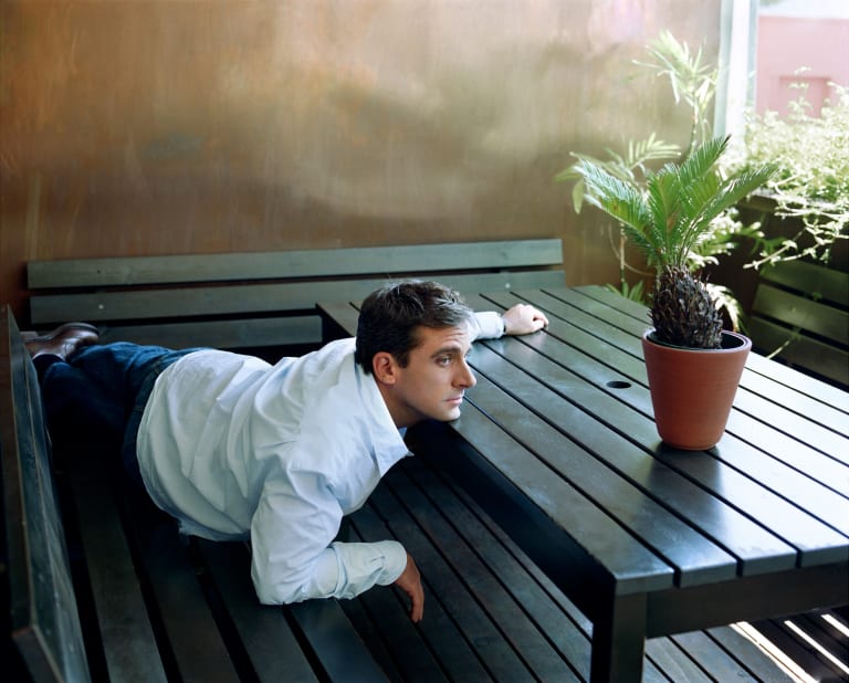 Portrait of comic actor Steve Carell. He is laying across a picnic table, looking off-camera to the right. There is a plant on the table next to him.