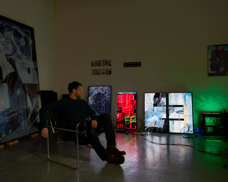 Image of the artist Chris Dorland Sitting in a chair looking to right at multiple cracked television monitors in his studio.