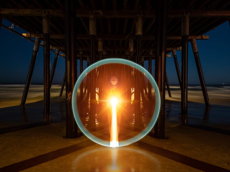 A color long exposure photograph of Russell Brown under a pier at night. He is pointing a light straight at the camera which is creating a flare in the shape of a perfect circle, framing his whole body.