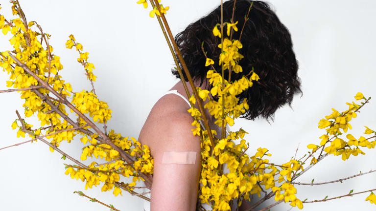 In this photo, a person in profile with bare arms, head lowered, holds under their arm a quiver of plant stems with yellow flowers.