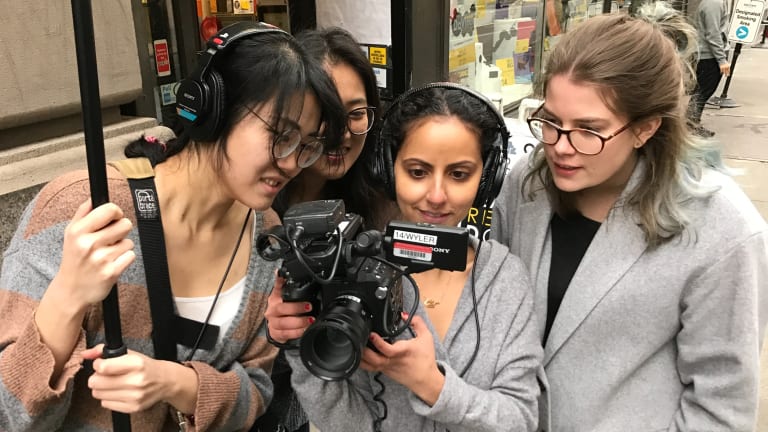 Four women wearing headphones stand outside and crowd around a camera, pointed at the viewer