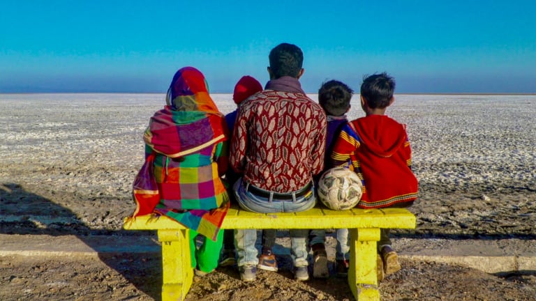 a family of two adults and three kids sits on a yellow bench with their back to the camera. They are brightly dressed, mostly in red, and stare across a flat white sand desert under a bright blue sky. A soccer ball is also on the bench.