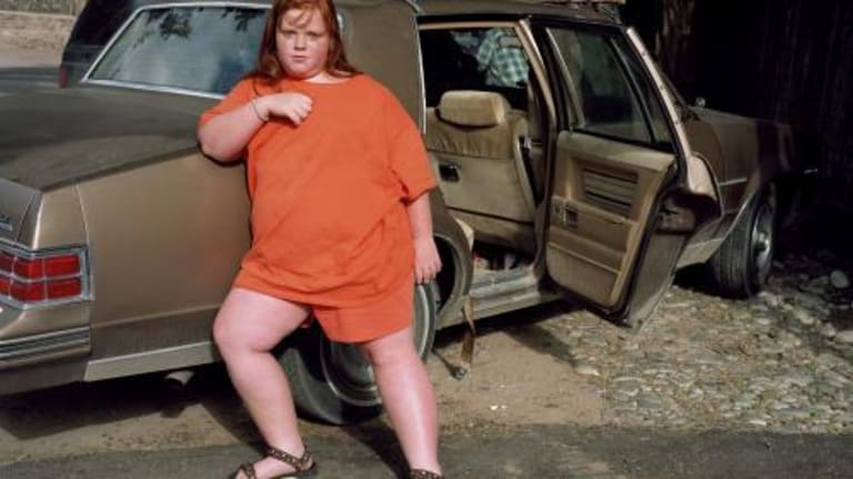 A person dressed all in orange is leaning against a brown vehicle with one back door open.