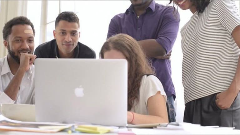 Five people are looking at the screen of an Apple laptop. They are all people of color (3 men, 1 woman) except one additional woman, the one typing on the computer, who is white. The man furthest left is African-American but has a Caucasian "flesh-toned" bandage on his forehead.