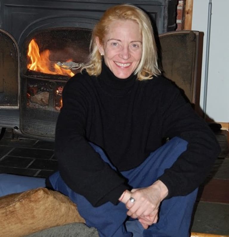 A blonde middle-aged woman in a black turtle neck and blue pants sits on a few pillows, smiling in front of a lit fireplace