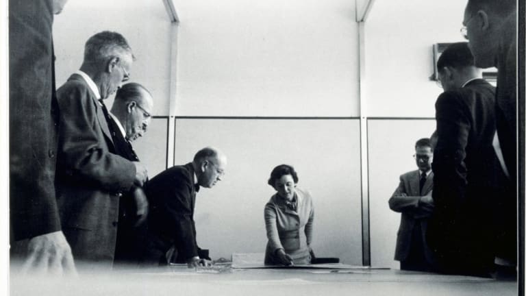 A black and white photograph down a conference table, with men on both sides of the table and a woman at the head, seeming to lead the conversation.