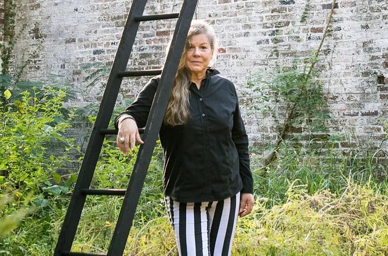 A woman stands next to a black ladder in a field in front of a white brick wall