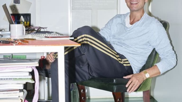 Photo of Kenny Schachter smiling and sitting at a desk with a large stack of magazines