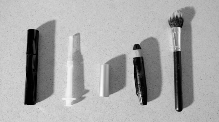 Black and white image of five objects placed side by side, including a medical syringe and several cosmetic applicators