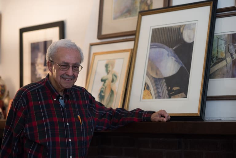 Richmond Jones wears a red plaid shirt and stands next to a fireplace with his hand resting on top of it. Above the fireplace are numerous framed paintings.