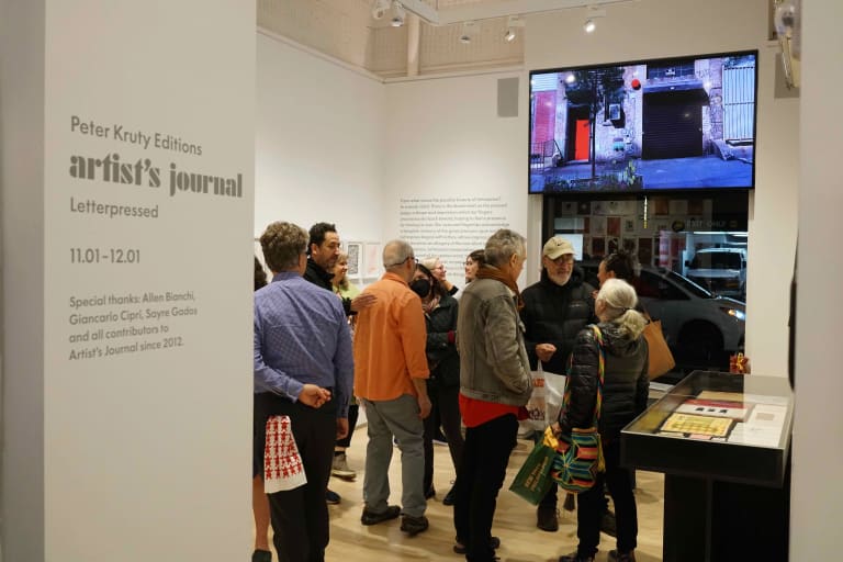 A view of an art exhibition reception from the entrance to a small gallery. There are about a dozen people in the room, a glass-topped display case against the right wall. The far wall has a large window with a video screen mounted directly above it. The exhibition information is in gray lettering on a narrow wall to the left side of the entrance.