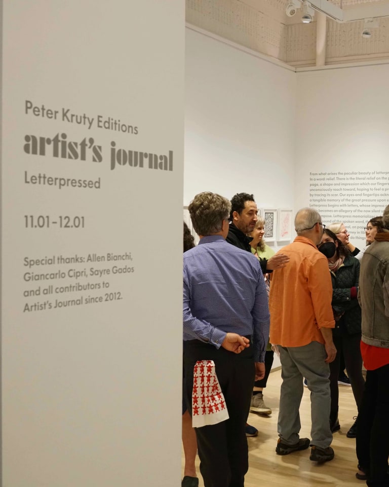 A view of an art exhibition reception from the entrance to a small gallery. There are about a dozen people in the room, a glass-topped display case against the right wall. The far wall has a large window with a video screen mounted directly above it. The exhibition information is in gray lettering on a narrow wall to the left side of the entrance.