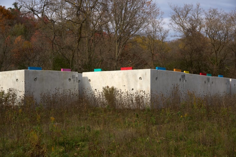 A photograph of a cement wall in a rural forest landscape. On top of the wall are multi-colored pieces of wood that make a repeating pattern.