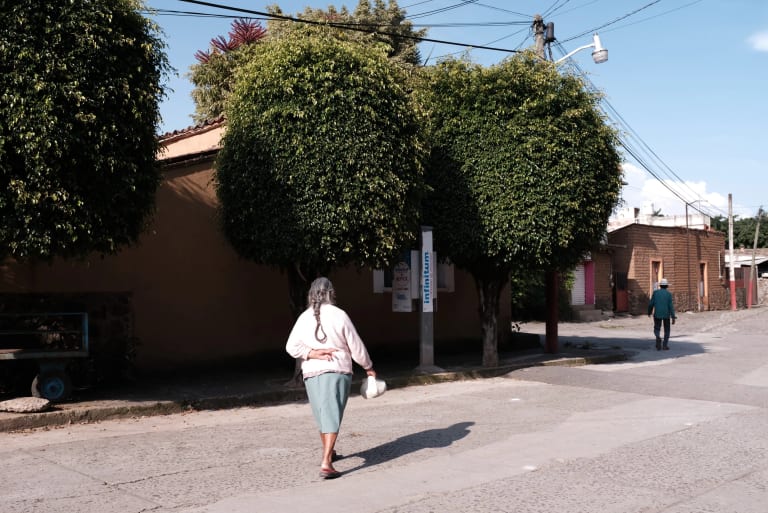 Woman walking with hand behind her back in a parking lot, following a man with a hat. In front of her is a building obscured by two trees.