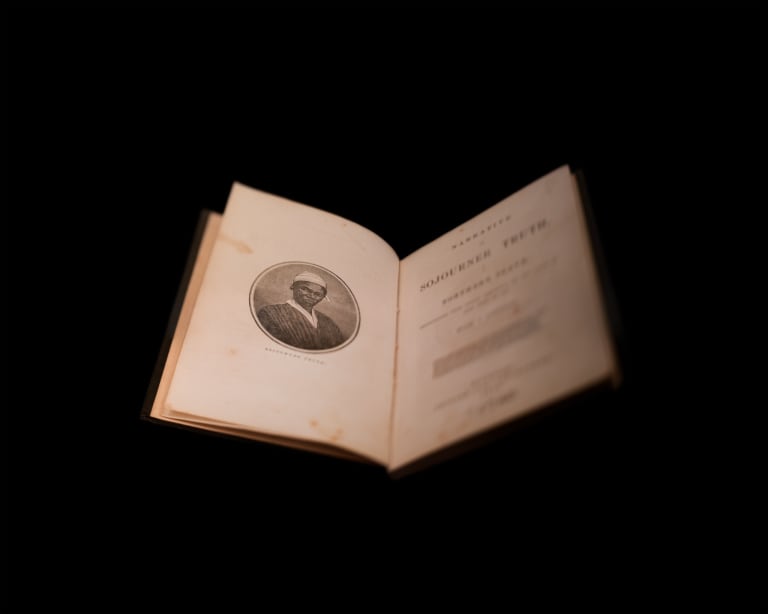 A staged setup of a historical book. It is photographed on a plain black background. The book is open to show an oval portrait of Sojourner Truth on the left page and the title and book info on the right page.