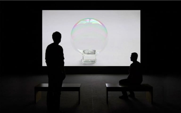 Two people in a room, looking at a screen that has a bubble sitting on a small cube.