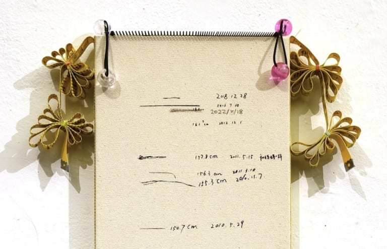 The top half of a sculpture made of a notebook with tassels and ribbons on the sides. Written on the notebook are different measurements.