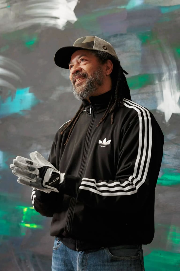 Portrait of a man, Gary Simmons, standing in front of a black wall with white, green and blue paint smudges and starts. The man is wearing a cap, an adidas jacket, and thick gloves.